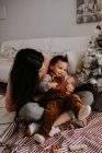 Von oben sieht man eine fröhliche junge Mutter in lässiger Kleidung, die ihren niedlichen lächelnden kleinen Sohn umarmt, während sie mit überkreuzten Beinen auf dem Boden neben dem geschmückten Weihnachtsbaum sitzt. — Stockfoto