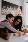 Улыбающаяся мать сидит за столом в украшенной гостиной и играет с милым маленьким сыном с деревянными игрушками — стоковое фото