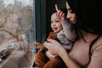 Side view of loving mother in casual clothes holding cute little son on hands while standing near window at home and pointing away — Stock Photo