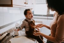Seitenansicht des fröhlichen kleinen Jungen in lässiger Kleidung mit Mehl auf den Wangen hilft Mutter beim Kochen von Gebäck in der modernen Küche — Stockfoto