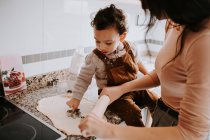 Vista lateral de niño alegre en ropa casual con harina en las mejillas ayudando a la madre a cocinar pastelería en la cocina moderna - foto de stock