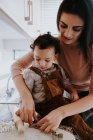 Vista laterale di allegra giovane madre in abiti casual preparare pasta con mattarello con figlio in piedi sulla sedia in cucina leggera — Foto stock