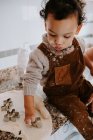 Niño lindo con el pelo rizado en ropa casual untado con galletas de harina de cocina con moldes de metal para hornear en la mesa en la cocina - foto de stock
