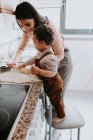 Vista lateral da jovem mãe alegre em roupas casuais preparando massa com rolo de pino com filho em pé na cadeira na cozinha leve — Fotografia de Stock