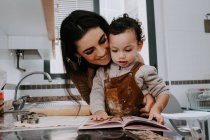Cheerful mother smiling and hugging son while reading recipe book on table during cooking delicious cookies in light kitchen at home — Stock Photo