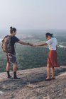Сверху вид на пару хипстеров в повседневной одежде, держащихся за руки и наслаждающихся закатом на каменном краю холма во время путешествия по Сигирии в Шри-Ланке — стоковое фото