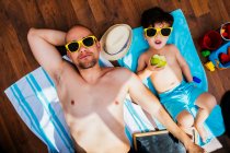Von oben sieht man einen fröhlichen Jungen mit gelber Sonnenbrille und blauer Badebekleidung, der lächelt, während er Vater Apfel füttert, der auf Handtüchern liegt und zu Hause am Strand liegt. — Stockfoto
