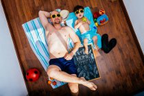 Сверху положительные отец и сын в желтых солнцезащитных очках и плавках лежат на полу на полотенцах и мечтают о летних каникулах на пляже во время карантина — стоковое фото