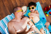 Desde arriba vista de niño alegre en gafas de sol amarillas y traje de baño azul sonriendo mientras se alimenta padre manzana acostado en toallas juntos tener playa en casa - foto de stock