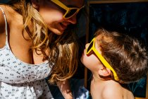 Visão lateral da mãe positiva em vestido branco e óculos de sol amarelos tocando narizes com filho nos mesmos óculos de sol na luz solar brilhante — Fotografia de Stock
