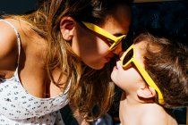 Visão lateral da mãe positiva em vestido branco e óculos de sol amarelos tocando narizes com filho nos mesmos óculos de sol na luz solar brilhante — Fotografia de Stock
