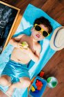 From above view of cheerful little boy in yellow sunglasses and blue swimwear eating apple while lying on towel on floor having home beach on quarantine — Stock Photo