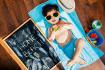 Сверху вид веселого маленького мальчика в желтых солнцезащитных очках и голубых купальниках, поедающего яблоко, лежа на полотенце на полу с домашним пляжем на карантине — стоковое фото