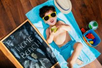 Сверху вид веселого маленького мальчика в желтых солнцезащитных очках и голубых купальниках, поедающего яблоко, лежа на полотенце на полу с домашним пляжем на карантине — стоковое фото