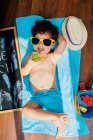 De cima vista de menino alegre em óculos de sol amarelos e azul swimwear comer maçã enquanto deitado em toalha no chão tendo praia em casa em quarentena — Fotografia de Stock