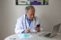 Anciano médico general masculino en vestido médico sentado en la mesa con el ordenador portátil en la clínica moderna y mirando en la pantalla del teléfono inteligente - foto de stock