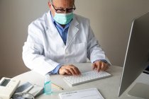 Médico senior con máscara facial y bata médica sentado en la mesa y tecleando en el teclado - foto de stock