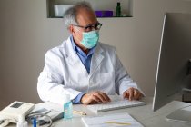 Médecin senior portant un masque facial et une blouse médicale assis à table et tapant sur le clavier — Photo de stock