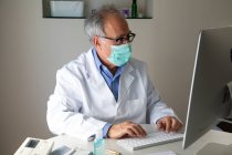 Médico sênior vestindo máscara facial e vestido médico sentado à mesa e digitando no teclado — Fotografia de Stock
