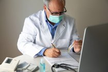 Médico masculino usando máscara protetora escrevendo no papel e olhando para o termômetro — Fotografia de Stock