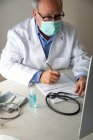 Médico geral do sexo masculino vestindo máscara protetora e vestido médico sentado à mesa e escrevendo relatório — Fotografia de Stock
