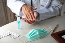 Чоловічий терапевт у медичній сукні сидить за столом у лікарні та омолоджує руки антисептиком — стокове фото