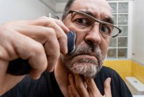 Middle aged bearded man in eyeglasses shaving with electric razor — Stock Photo