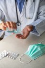 Мужской терапевт в медицинском халате сидит за столом в больнице и дезинфицирует руки антисептиком — стоковое фото