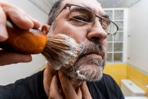 Homem aplicando espuma de barbear com escova enquanto se prepara para o procedimento de barbear no banheiro — Fotografia de Stock