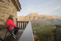 Vue latérale de la fille en vêtements décontractés assis sur une chaise sur la terrasse en bois de la maison en pierre en Espagne et admirant paysage de montagne pittoresque tout en regardant loin — Photo de stock