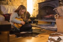 Focused teen ragazza in abiti casual seduta con gambe incrociate su comodo divano e giocare ukulele in casa di pietra durante le vacanze in Cantabria — Foto stock