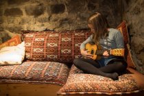 Focalisé adolescent fille en vêtements décontractés assis avec les jambes croisées sur canapé confortable et jouer ukulele dans la maison en pierre pendant les vacances en Cantabrie — Photo de stock