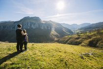 Back view of anonymous female hiker embracing kids while standing on green meadow and observing picturesque landscape of mountains on sunny day in Cantabria — Stock Photo