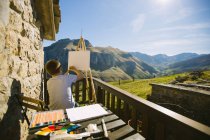 Back view of faceless boy in white t shirt sitting at easel on picturesque veranda of country house and drawing picture in Cantabria — Stock Photo