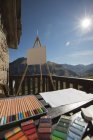 Набор разноцветных карандашей и различных инструментов для рисования на деревянном столе возле мольберта с холстом на солнечной террасе старинного каменного дома в Испании — стоковое фото