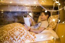 Контент мальчик лежит в уютной кровати и с помощью планшета во время отдыха в спальне украшен светящимися уютными гирляндами — стоковое фото
