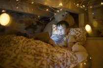 Контент мальчик лежит в уютной кровати и с помощью планшета во время отдыха в спальне украшен светящимися уютными гирляндами — стоковое фото