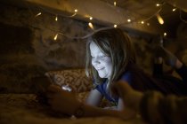 Очаровательная девушка-подросток в повседневной одежде отдыхает на уютной кровати под светящейся гирляндой и просматривает планшет в свободное время — стоковое фото
