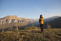 Back view of faceless girl in casual clothes standing alone on green hill and enjoying picturesque view of mountains and sky in Cantabria — Stock Photo