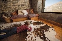 Niño acostado en el suelo sobre una alfombra acogedora y dibujo con lápices de colores en cuaderno de bocetos escalofriante acogedor salón de casa de piedra en Cantabria - foto de stock