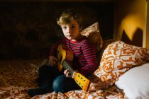 Niño concentrado en traje casual sentado en una cómoda cama en el dormitorio y jugando ukelele mientras se enfría en casa en Cantabria - foto de stock