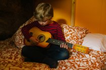 Concentrated child in casual outfit sitting on comfortable bed in bedroom and playing ukulele while chilling at home in Cantabria — Stock Photo
