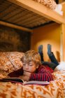 Enfant rêveur en tenue décontractée couché sur un lit confortable et profitant de conte de fées intéressant dans le livre d'histoire pour enfants — Photo de stock