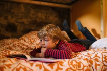 Мечтательный ребенок в повседневной одежде лежит на уютной кровати и наслаждается интересной сказкой в книге рассказов для детей — стоковое фото
