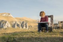 Adorable child sitting on wooden bench and painting on canvas during weekend on background of magnificent mountainous landscape — Stock Photo