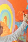Chica rubia creativa en ropa casual sentado en el alféizar de la ventana contra la ventana y la pintura con pincel grande arco iris multicolor sobre lienzo naranja - foto de stock