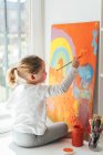 Ragazza bionda creativa in abiti casual seduta sul davanzale della finestra contro la finestra e pittura con pennello grande arcobaleno multicolore su tela arancione — Foto stock