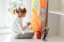 Креативна блондинка в повсякденному одязі сидить на підвіконні проти вікна і малює пензлем велику різнокольорову веселку на помаранчевому полотні — стокове фото