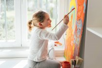 Творческая блондинка в повседневной одежде сидит на подоконнике у окна и рисует кисточкой большой разноцветной радугой на оранжевом холсте — стоковое фото