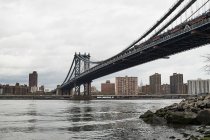De baixo de Manhattan Bridge visto da margem do rio com edifícios da cidade e céu cinza nublado em segundo plano na cidade de Nova York — Fotografia de Stock